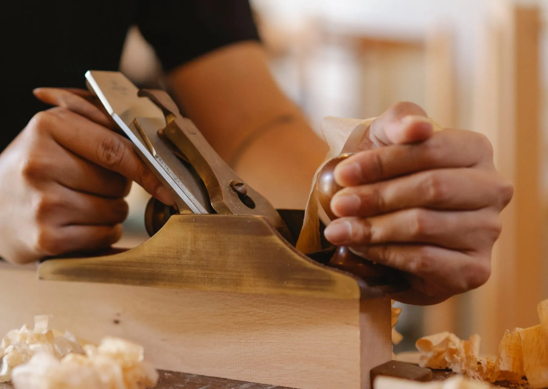 Hands planing a pale timber board, with shavings on the workbench