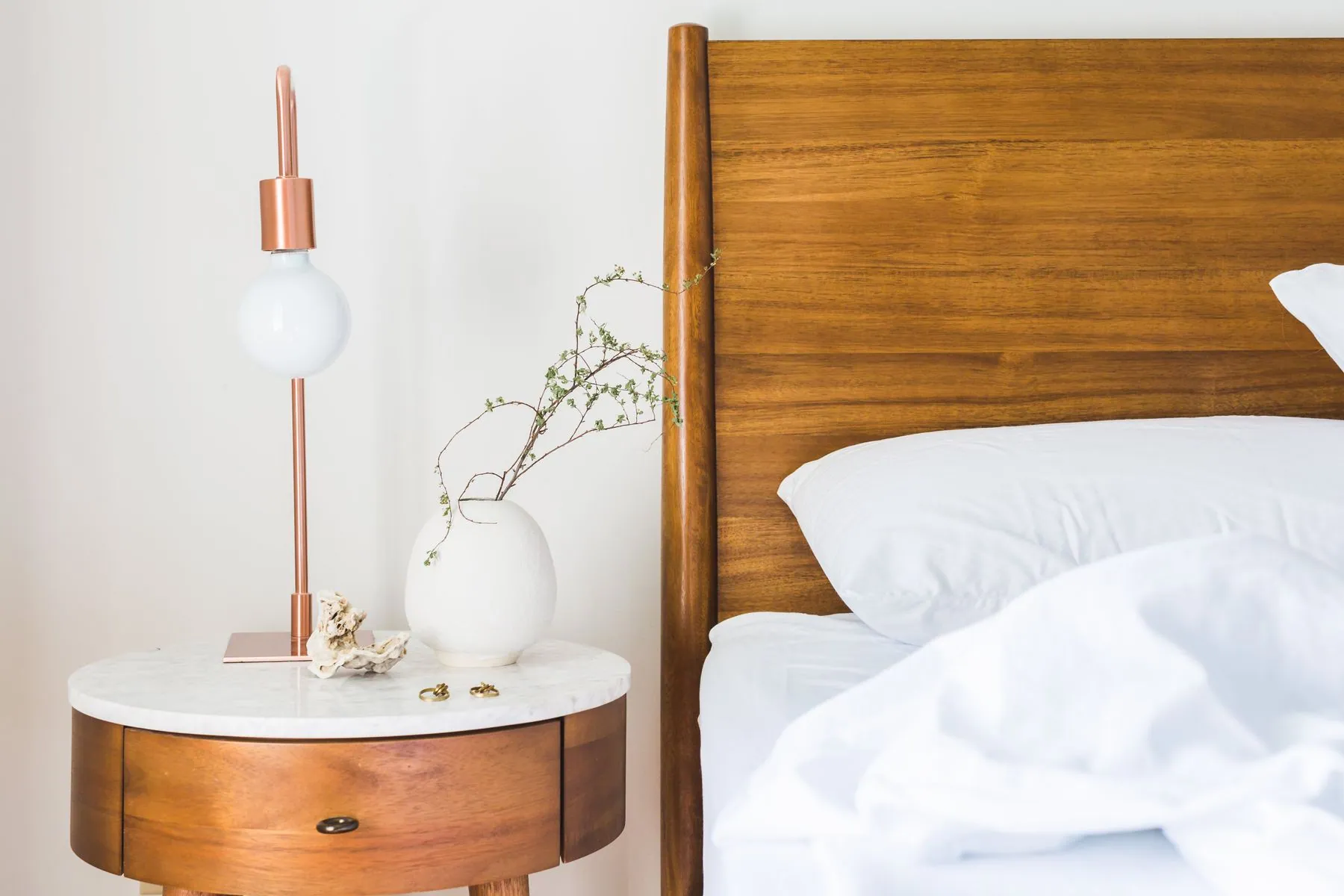 Round bedside table with a lamp beside a walnut headboard and white bedding