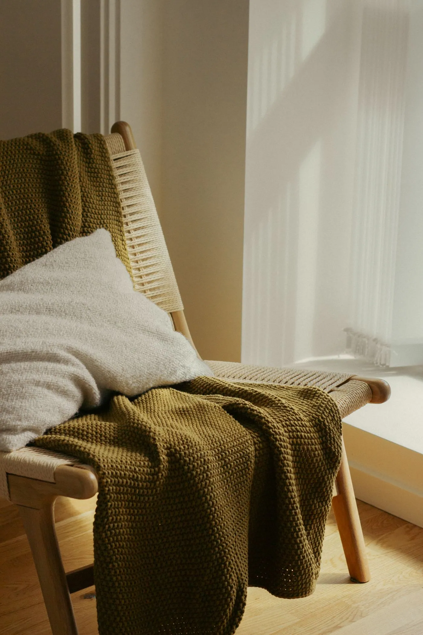 Woven wooden armchair with a green knitted blanket and a cushion beside a sunny window