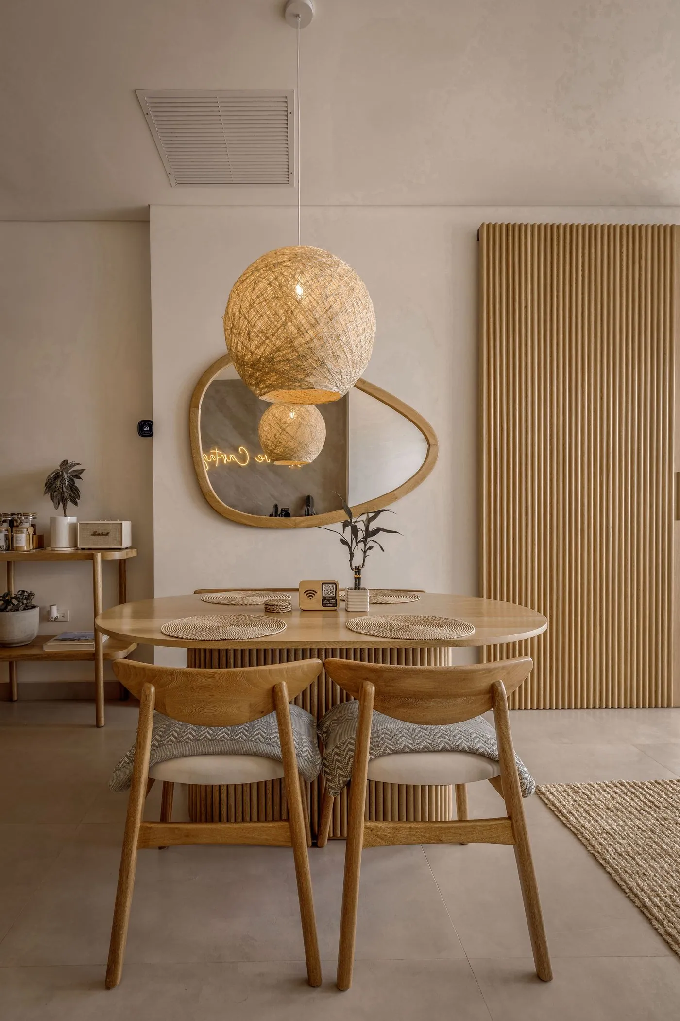 Round wooden dining table under a woven pendant light, with a slatted wood wall